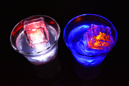A champagne glass with a pattern of branches and leaves filled with a white drink is located on a black matte background. Small ice cubes glowing in different colors float in a glass, party, celebratiの写真素材