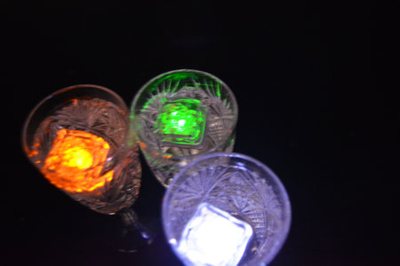 Crystal champagne glasses with carvings in the form of snowflakes are located on a black matte background. Bright multicolor doidic glowing ice cubes float in the drink.の写真素材