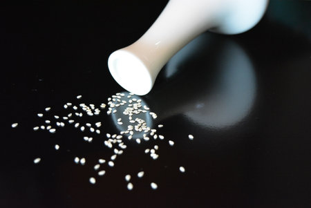 White sesame seeds spilled from a white mortar on a black background. Home furnishings, style and food, white ceramic vase with white sesame seeds set on black glossy background.の写真素材
