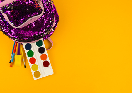 Stationery items from a backpack on a yellow background. Purple girls briefcase with sequins shine....