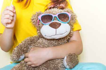 The child is holding a favorite toy bear. Sweet lollipop. Little girl in a yellow T-shirt and glasses.の写真素材