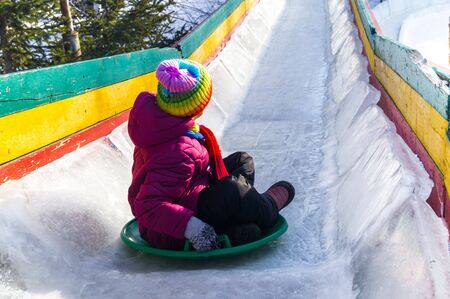 Child rolls down an ice slide. Merry winter sport family. Bright clothes rainbow hat.