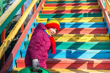 Child holds a sled goes down the ice slide. Merry winter sport family. Bright clothes rainbow hat.の写真素材