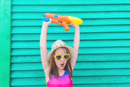 Girl with a water gun. Baby in hat and glasses on a wooden background. Active holidays with family...