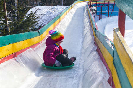 Child rolls down an ice slide. Merry winter sport family. Bright clothes rainbow hat.の写真素材