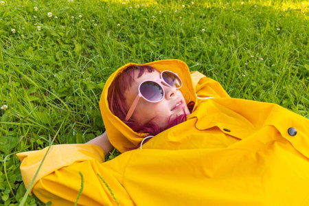Girl in a yellow raincoat. Outdoors walk. Kid lies on the green grass. Hipster child teenager in pink glasses. Summer heat.の写真素材