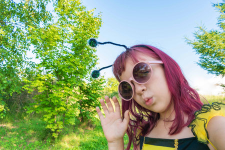 Girl in a bee costume with pink hair outdoors. Looks into the camera takes a selfie Child pretends to fly. Blue sky backgroundの写真素材