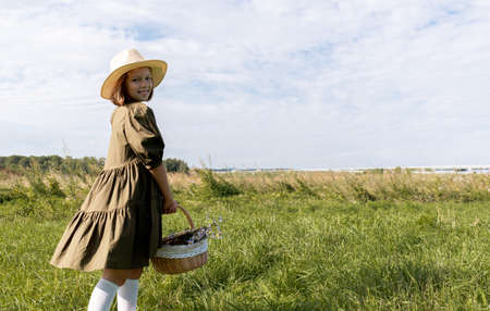 Girl in a linen dress and a straw hat walks on a flower field. Wicker basket in hands. Wellness and freedom concept.の写真素材