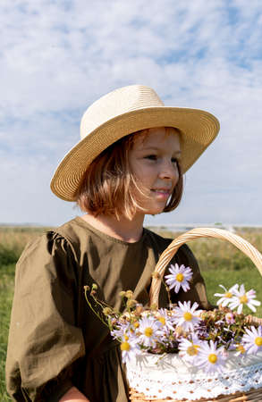 Girl in linen dress and a straw hat stands with a basket of daisies. Glade with flowers. Wellness and freedom concept.の写真素材