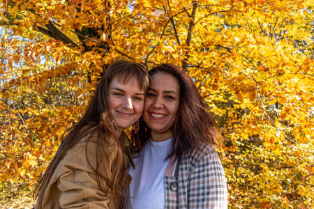 Two diverse female friends are chatting and hugging while standing in autumn forest. female friendship. outdoors. Cheerful person.の写真素材