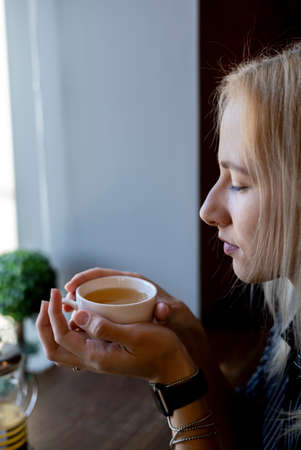Young adult woman holds a white cup with a hot drink in her hands. teatime. Green tonic herbal tea with antioxidantsの写真素材