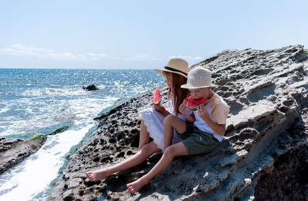 Boy and a girl in beach clothes are sitting on a rock near the sea and eating watermelon. Brother...