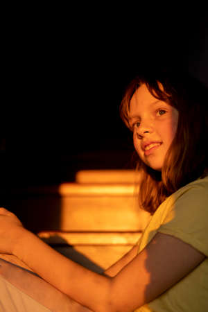 Young teen girl is sitting on the stairs with light and shadows from shutters or blinds. Little girl...
