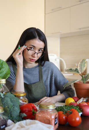 Young Asian woman writes down recipe for a dish in notebook. Zero waste. Vegetarian woman. Conscious consumption. fresh vegetables and fruitsの写真素材
