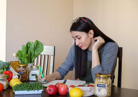 Young Asian woman writes down recipe for a dish in notebook. Zero waste. Vegetarian woman. Conscious consumption. fresh vegetables and fruitsの写真素材