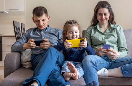 Family sit on the couch and hang out on their phones. Mom dad and daughter spend time with gadgets....