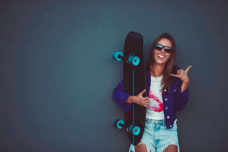 Young happy girl in sunglasses showing shaka with skateboard in front of gray wall.の写真素材
