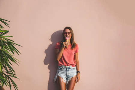 Model girl in jeans shorts and pink t-shirt licking ice cream on pink background