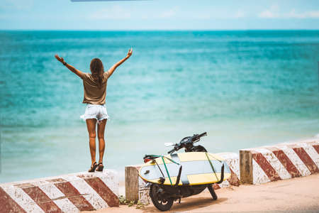 Happy young woman enjoying life and feeling free with open arms in summer vacation travel at the sunny day on the beach near motorbike with longboard surfboard. Freedom and carefree lifestyle conceptの写真素材