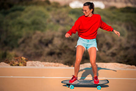 Sporty carefree woman in jeans shorts, red sneakers and hoodie riding surf skateboard in sunny...