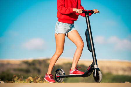 Fit teenage girl in red hoodie and blue denim shorts holding handlebar of electric scooter on the road. Summer outdoor activities.の写真素材