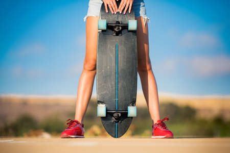 Stylish fit and sporty woman with beautiful legs and in red sneakers holds skateboard between her legs on shiny summer day . Outdoor activities. Street culture.の写真素材