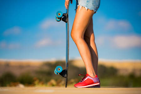 Stylish fit and sporty woman with beautiful tanned legs and in red sneakers holds longboard on shiny summer day . Outdoor activities. Street culture.の写真素材