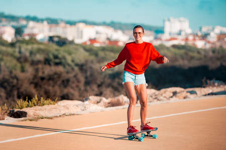 Sporty cheerful smiling woman in blue shorts, red sneakers and hoodie riding skateboard in sunny summer day. Vacation activitiesの写真素材