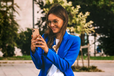 Happy smiling businesswoman in blue blazer using smartphone. Video call. Talking with friends on cell phone. Typing message or shop online or checking mobile application or social mediaの写真素材