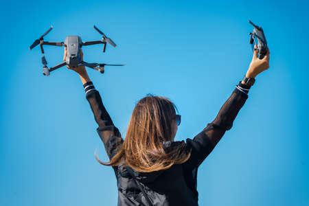 Young woman in black jacket holding remote control and drone before the flight on the blue sky...