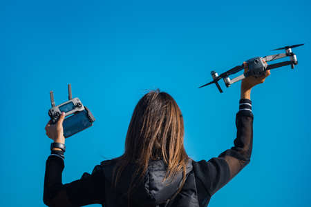 A back side view of young woman in black jacket holding remote control and drone before the flight on the blue sky background. Flying quadcopter outside in summer.の写真素材
