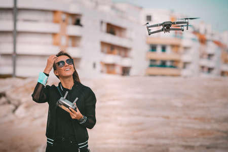 Cute modern smiling lady pilot in dark stylish clothes controls the drone with remote control. Outdoors hobby.の写真素材