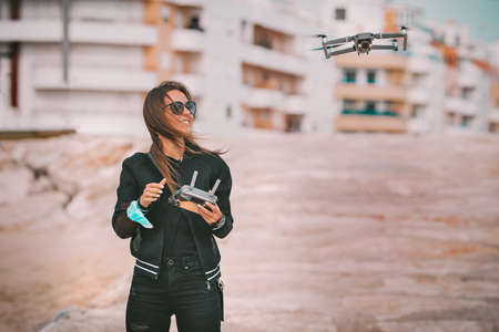 Pretty modern smiling woman pilot in dark stylish clothes flying drone with strong wind in city. Fly drone with permission. Outdoors hobby.の写真素材