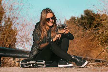 Young modern woman in stylish dark clothes and sunglasses preparing to start flying drone on sunset or sunrise.の写真素材