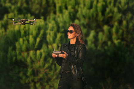 Young smiling modern woman in dark stylish clothes operating the drone by remote control in forest at sunset.の写真素材