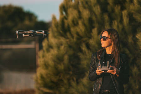 Young modern smiling woman pilot in dark stylish clothes operating the drone by remote control. Flying drone in forest at sunset. Female launches a drone for flight in the evening.の写真素材