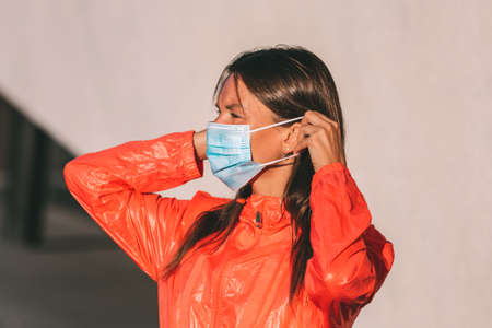 Beautiful young woman in orange clothes puts on or takes off a disposable medical mask. Protection against coronavirus.の写真素材