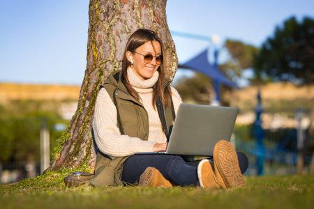 Woman with headphones and laptop sitting on the grass in the park. Freelance work. Work or study online. New tech usage conceptの写真素材