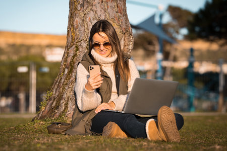 Photo of happy caucasian woman sitting on the grass outdoors in the park using laptop and mobile phone. Online and freelance work conceptの写真素材