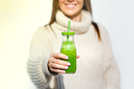 Smiling woman in woollen sweater holding bottle with green homemade detox smoothie or juice. Healthy...