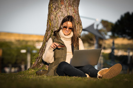 Happy smiling woman sitting outdoors in the park. Holding credit card and using laptop computer and making an online payment for a purchase. Online shoppingの写真素材