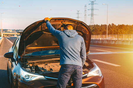 Man with his broken car checking engine on the highway roadside at sunset. Automobile breaks down on the autobahn. Open the car bonnetの写真素材