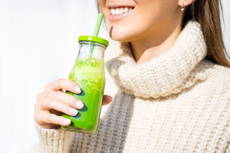 Smiling woman in woollen sweater drinking green homemade detox smoothie or juice. Healthy eatingの写真素材