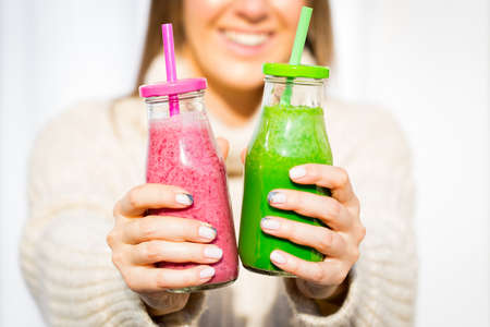 Happy smiling woman in woollen sweater giving 2 bottles of pink and green fresh smoothieの写真素材