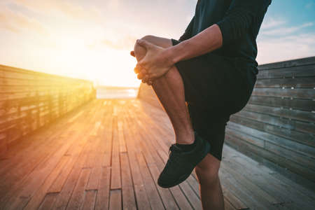 Healthy young man warming up and stretching legs before workout outdoors at sunset or sunrise. Stretching gluteus maximus muscle. Runner doing fitness stretching exercises before runの写真素材