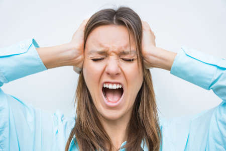 Portrait of young woman with wide open mouth, closed eyes and hands on head screaming or shouting. Concerned and stressed woman in panicの写真素材