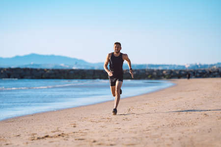 Runner in motion. Young sporty athletic man in black sportswear running or jogging along the beach near the ocean. Healthy lifestyleの写真素材