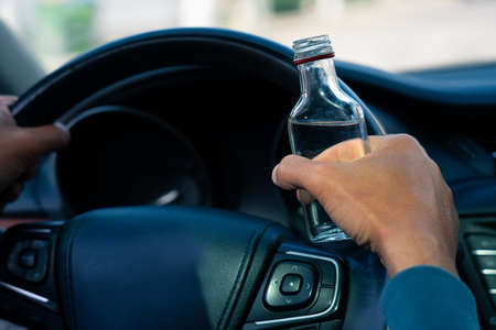 Closeup of drunk man hands on the steering wheel with a bottle of vodka. Driving under alcohol influence. Drunkenness. Dangerous driving conceptの写真素材