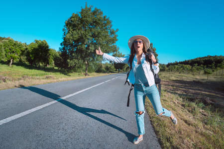 Young attractive woman tourist with backpack hitchhiking on the road. Traveling by autostop. Backpacker stopping car with thumbs up. Picking up hitchhikers. Summer vacation adventure. Ready to travelの写真素材