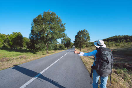 Young woman with backpack travelling by car. Hitchhiking on the road. Traveling by autostop. Backpacker stopping car with thumbs up. Way to adventure. Budget or cheap travel.の写真素材